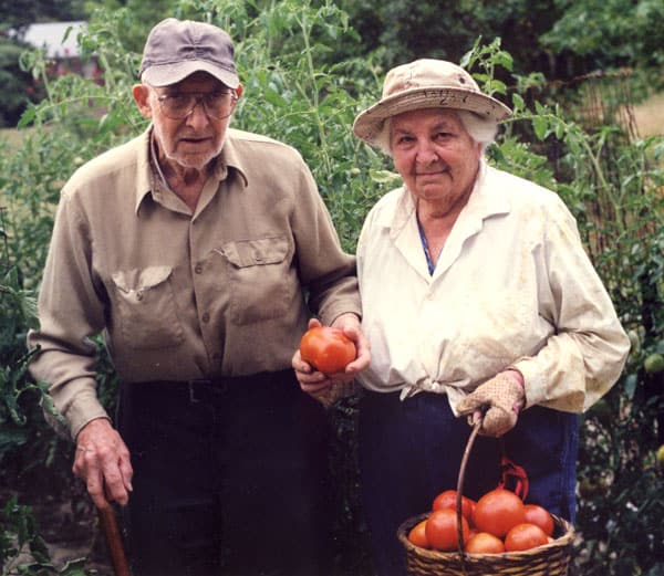 Married couple (Paul and Anna Atkins) in hats and collared shirts holding a basket of ripe red tomatoes